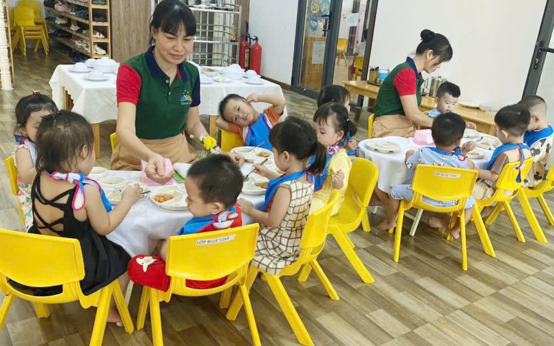 Các bé ăn trưa ở bàn tròn, cô đi quanh chia thức ăn