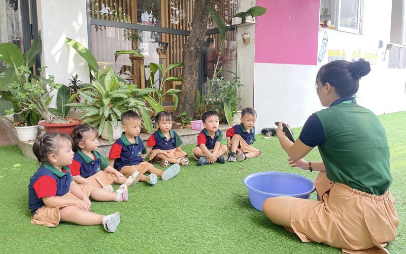 Cô ngồi trên cỏ hướng dẫn các bé chơi với chậu nước