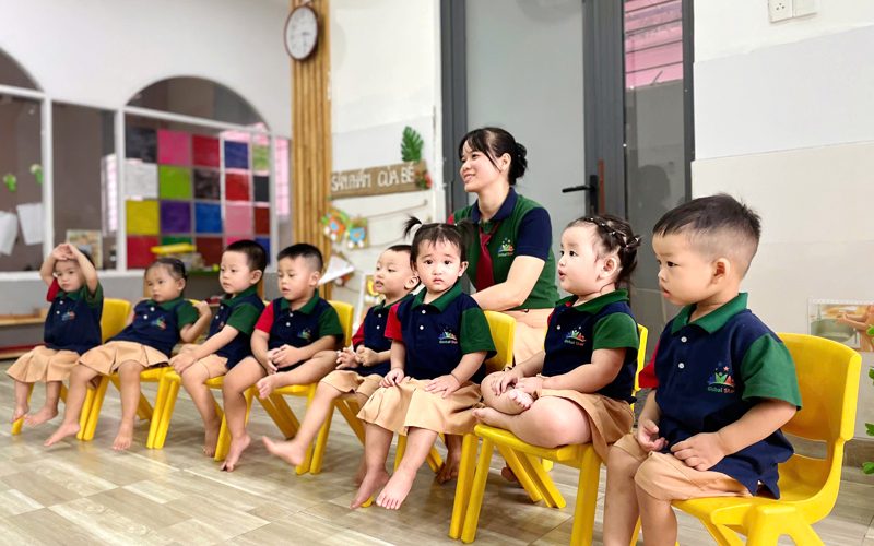 Các bé mặc đồng phục ngồi ghế vàng thành hàng trong lớp, cô ngồi phía sau