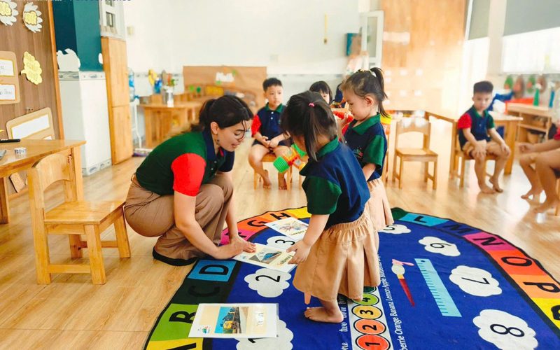 Cô ngồi xuống ngang tầm bé trên thảm chữ số trong lớp